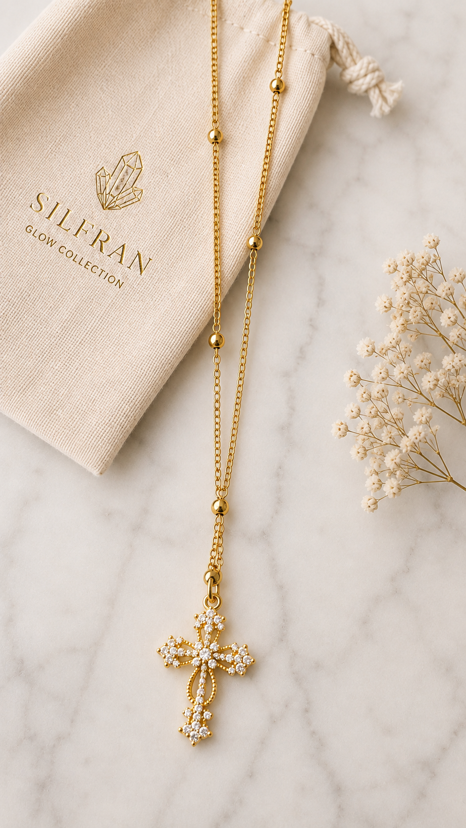A gold cross pendant necklace with clear crystals, displayed on a white marble surface next to a beige drawstring bag printed with 'SILFRAN GLORY COLLECTION' and dried baby's breath flowers.