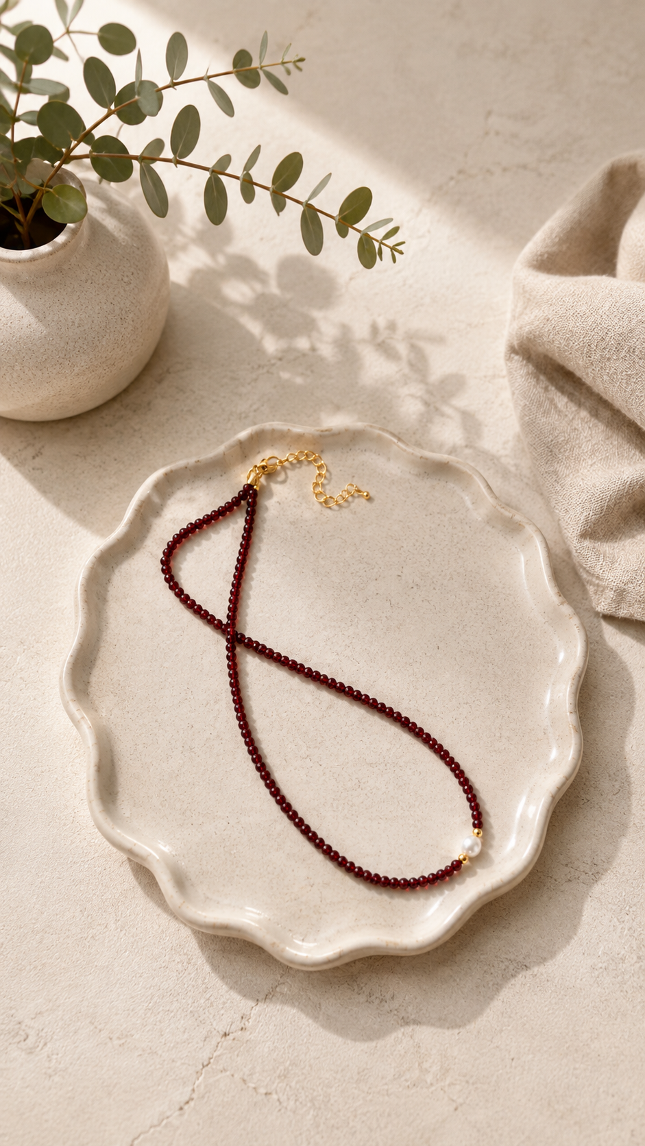 A beaded necklace with dark red beads and a gold clasp, displayed on a scalloped cream ceramic plate with a small plant and beige fabric in the background.