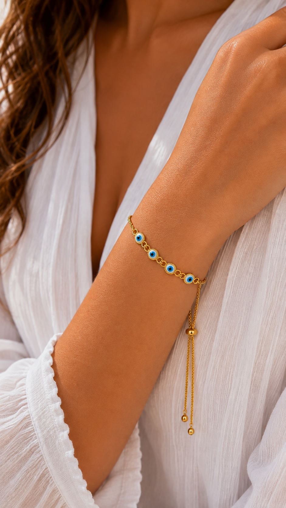 A gold lariat-style bracelet with small blue and white enamel evil eye charms, worn on a woman's wrist over a white blouse.