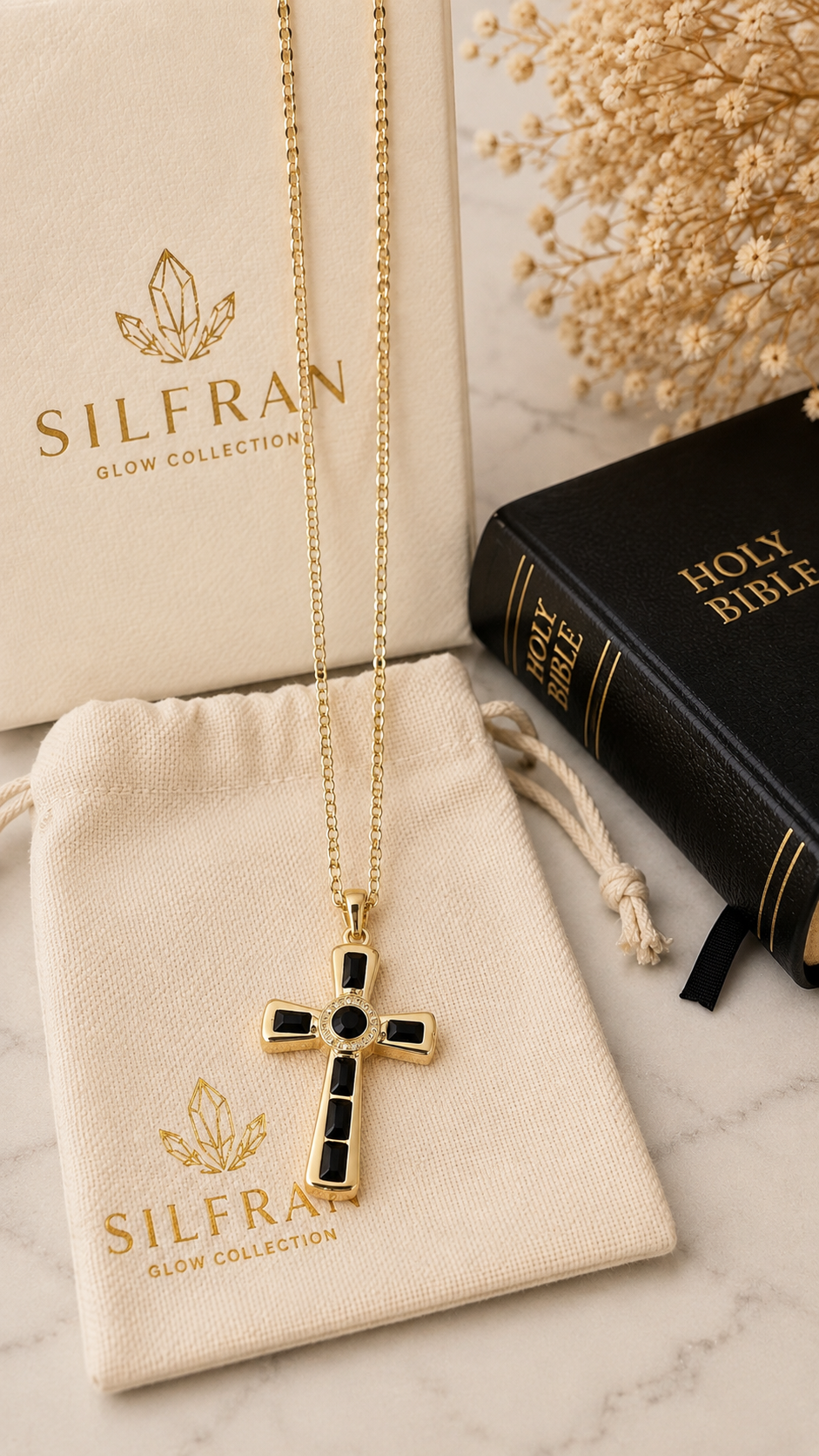 A gold cross pendant necklace with black inlay details from the Silfran Glow Collection, displayed on a cream-colored drawstring pouch next to its branded box and a black Holy Bible on a marble surface.