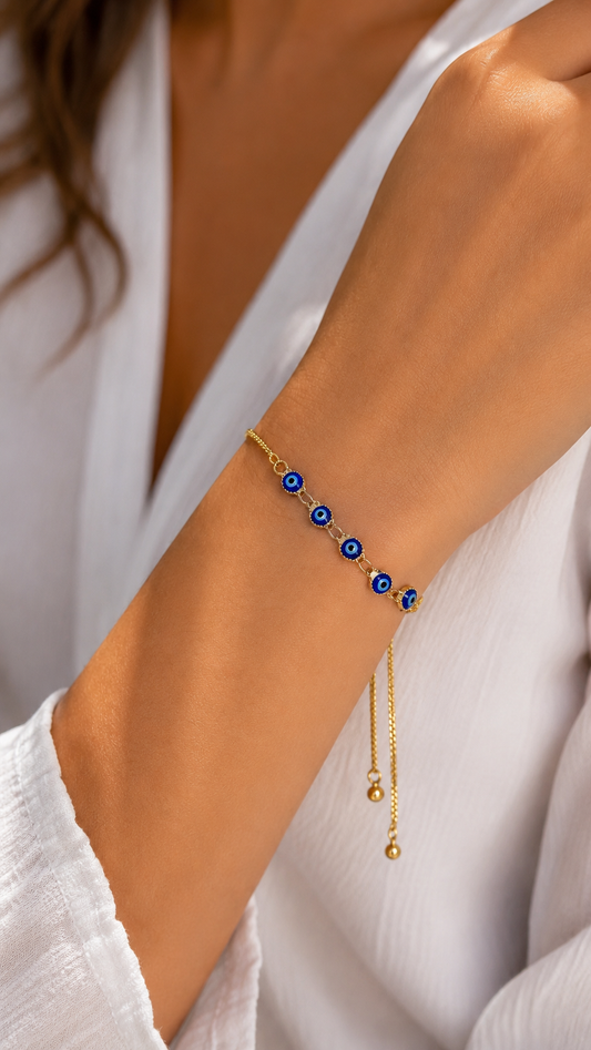 A gold chain bracelet with five blue and white evil eye charms worn on a woman's wrist, shown in a close-up lifestyle shot.