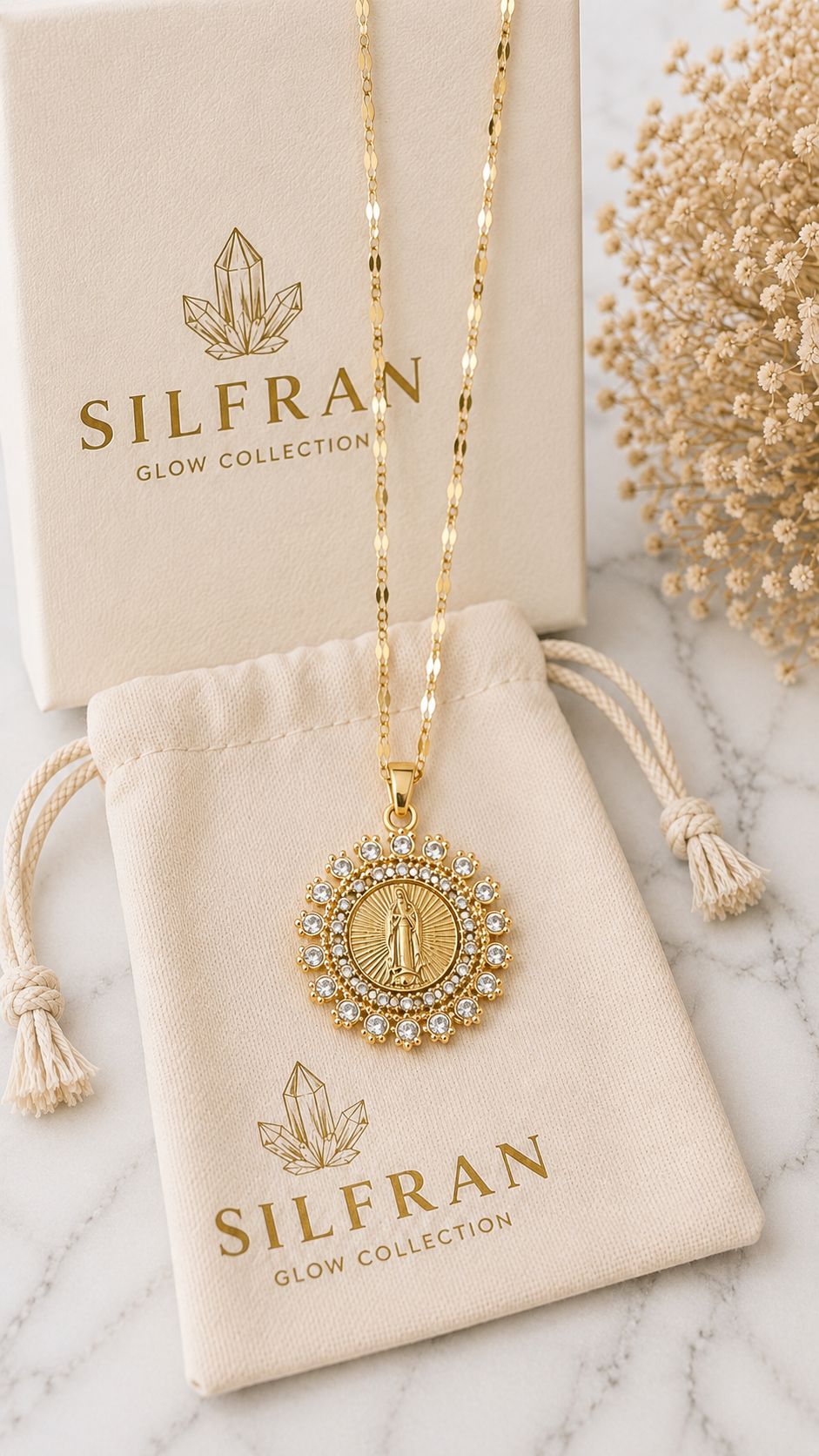 A gold-toned SILFRAN pendant necklace with a circular medallion, displayed on a cream drawstring pouch and matching box on a light marble surface with dried floral decor.