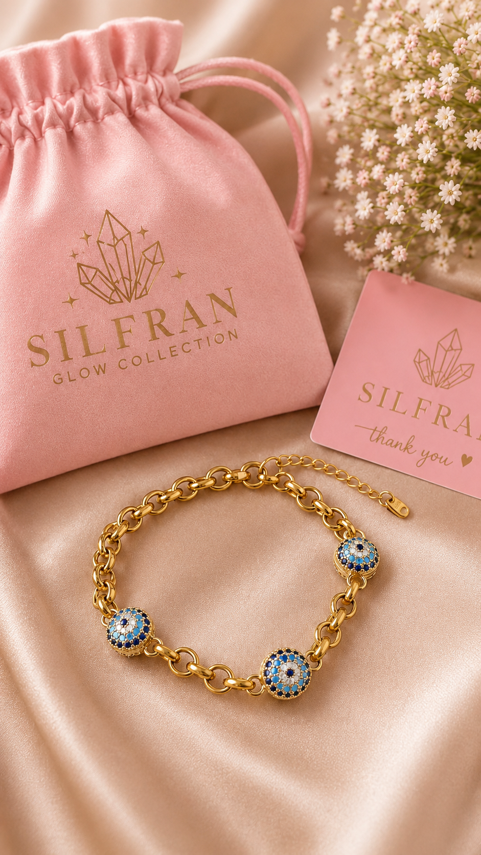 A gold chain bracelet with three blue and white evil eye charms, displayed on a beige satin fabric next to a pink drawstring pouch and a pink thank you card from the Silfran Glow Collection.