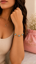 A close-up lifestyle shot of a model wearing a gold chain bracelet with blue and white evil eye charms. The model is wearing a beige ribbed tank top, and a pink drawstring bag with the brand name 'SILERAN' is visible in the background next to dried flowers.