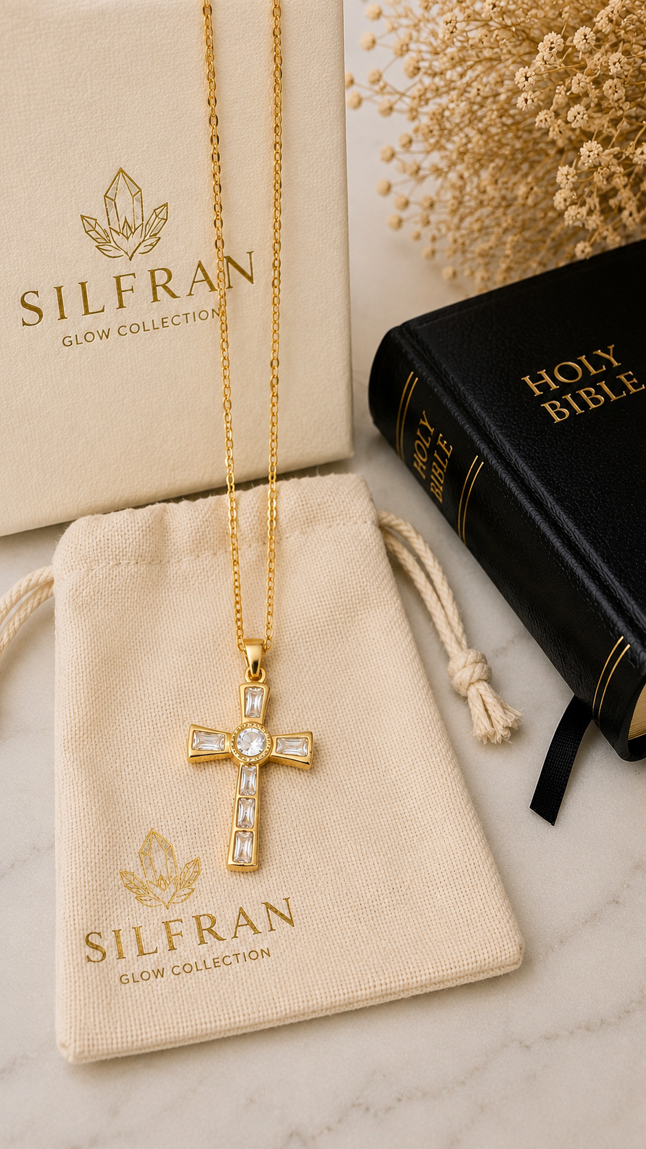 A gold cross pendant necklace with clear crystals, displayed on a cream drawstring pouch branded SILFRAN, with a matching branded box and a black Holy Bible on a light marble surface.