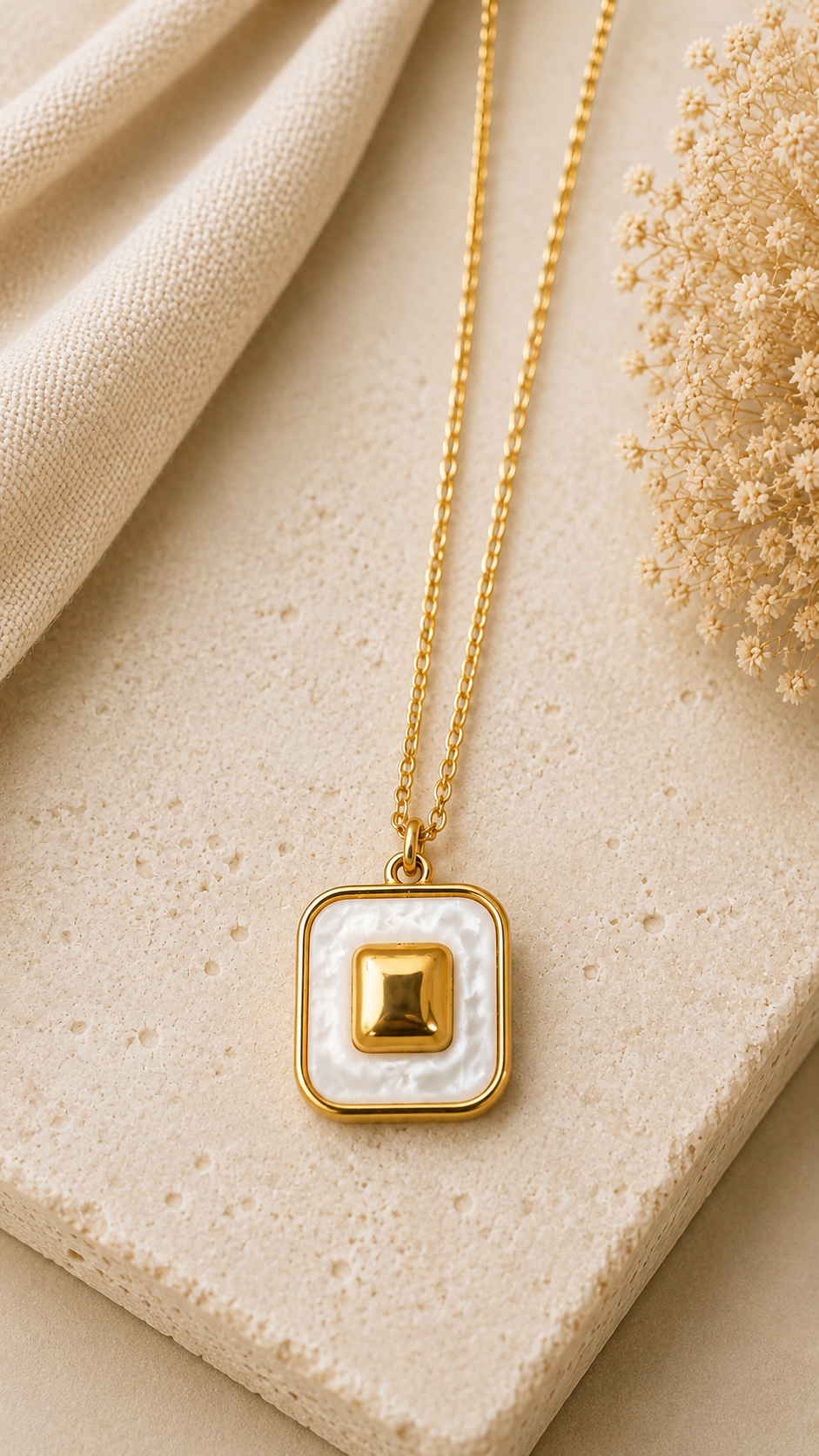 A gold square pendant necklace with a white textured center and a small gold square accent, displayed on a beige stone surface with a cream fabric and dried floral prop.