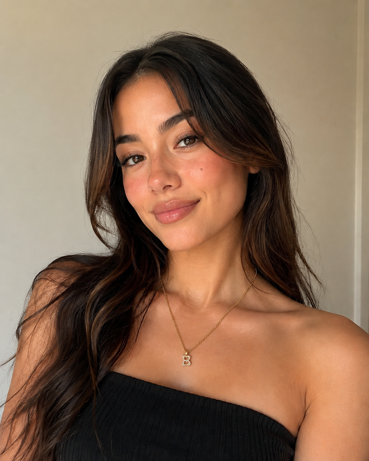 Woman with long dark hair wearing a black strapless top and a necklace with a 'B' pendant.