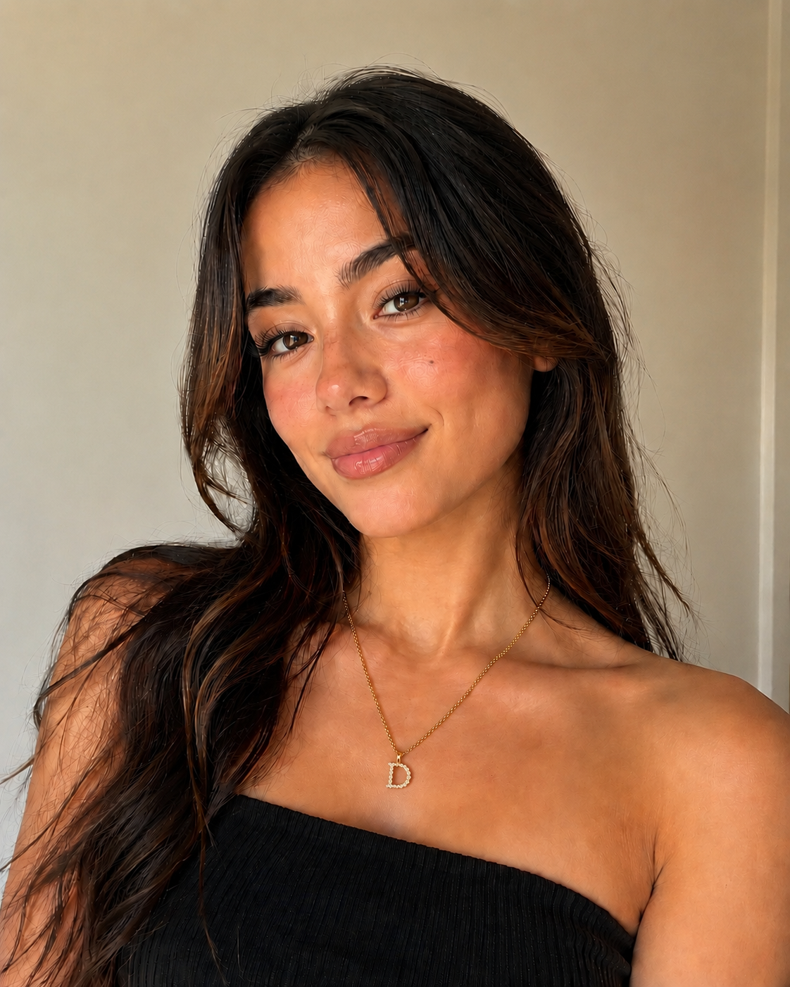 Woman wearing a black strapless top with a necklace featuring a 'D' pendant against a neutral background.