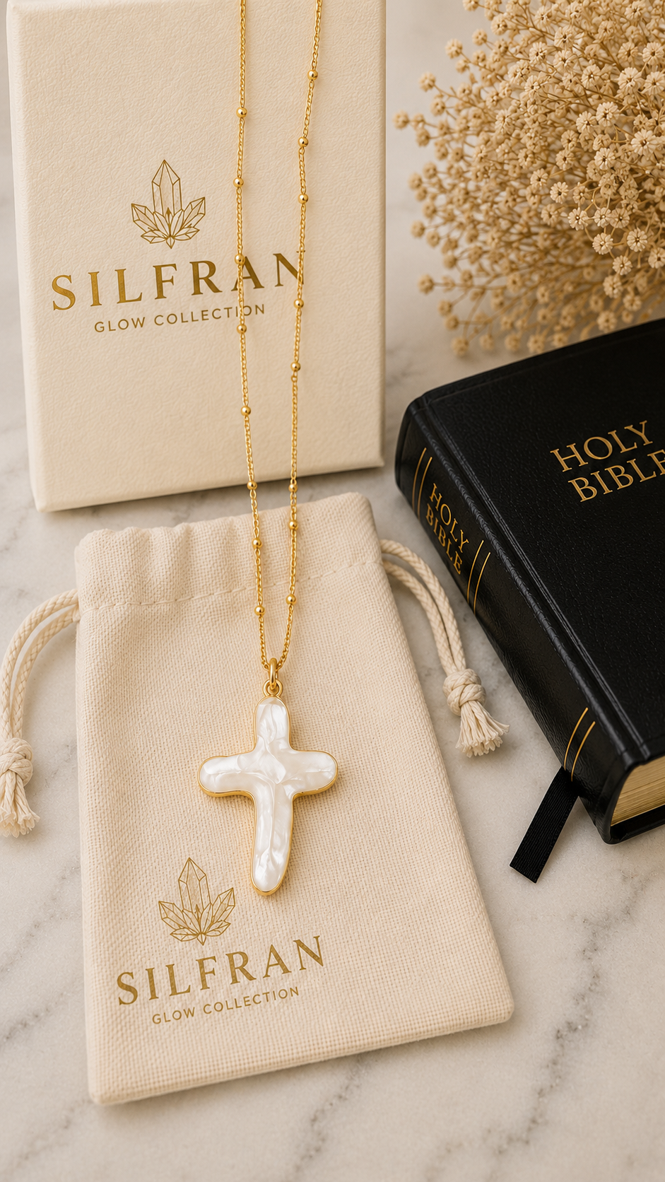 A gold chain necklace with a white mother-of-pearl cross pendant from the Silfran Glow Collection, displayed on a cream drawstring pouch next to its branded box and a black Holy Bible on a marble surface.