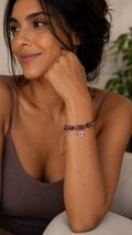 A woman wearing a delicate gold chain bracelet with a small pink arrow charm and purple beads on her wrist, posing in a ribbed brown camisole.