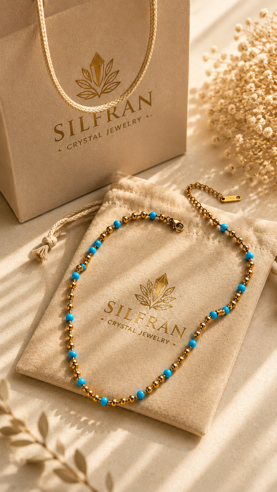 A beaded necklace with turquoise blue and gold beads is displayed on a beige velvet jewelry pouch branded SILFRAN, with a matching branded shopping bag and dried floral props in a warm, sunlit setting.