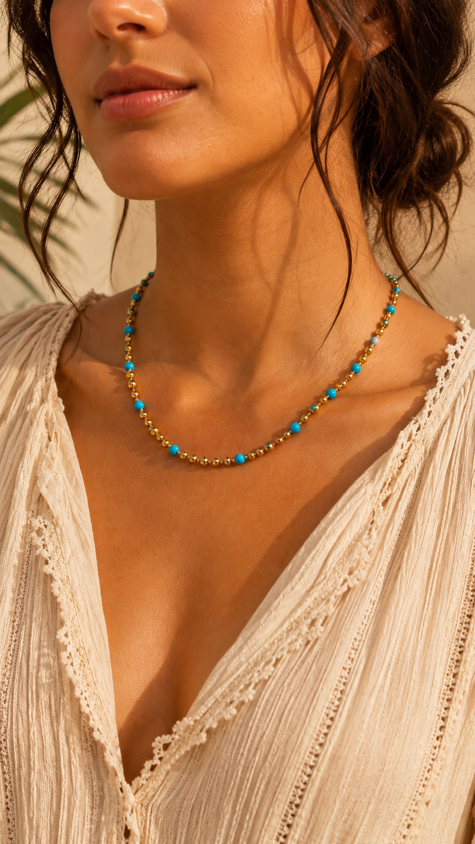 A close-up lifestyle shot of a woman wearing a delicate beaded necklace with turquoise blue and gold beads, paired with a cream-colored lace-trimmed blouse.