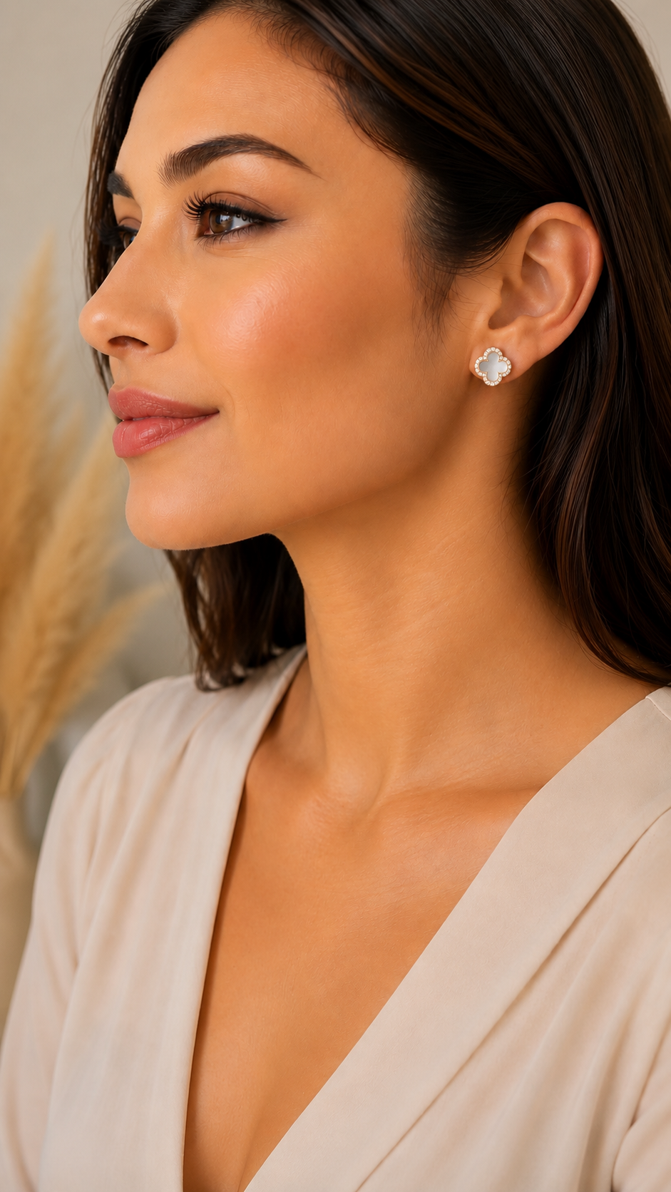 A close-up side profile of a woman wearing a small, clear, flower-shaped stud earring. She has dark brown hair and is wearing a light beige V-neck top, set against a soft, warm neutral background with dried pampas grass on the left.