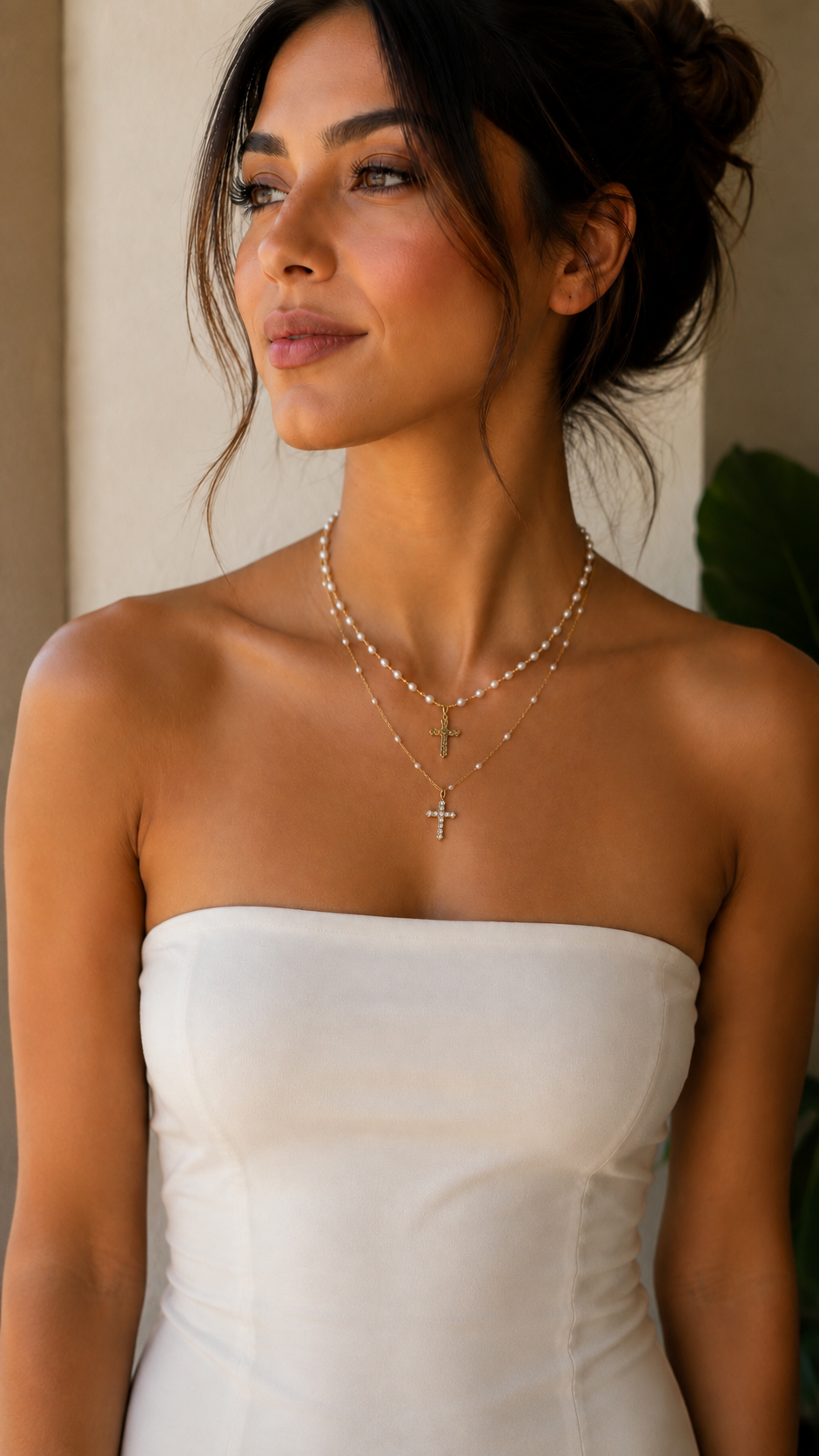 A woman wearing a delicate layered necklace with a small cross pendant, styled with a white strapless dress in a warm indoor setting.